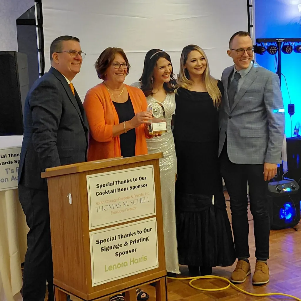 Five smiling people, one holding an award, stand next to a podium at an event.