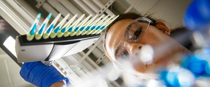 Scientist in safety glasses and blue gloves looks up at pipette tips with blue liquid.