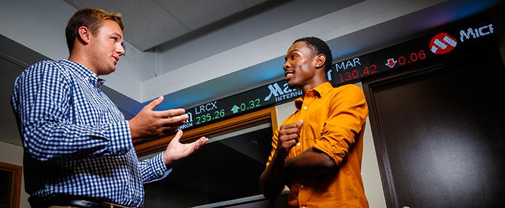 Two people talking in front of a stock market display.