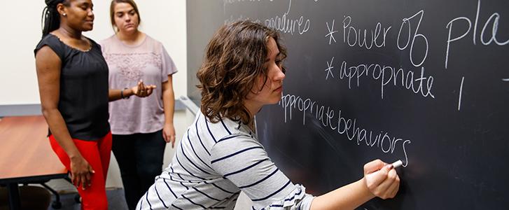 Three people in a classroom, one writes on a blackboard while two others watch.