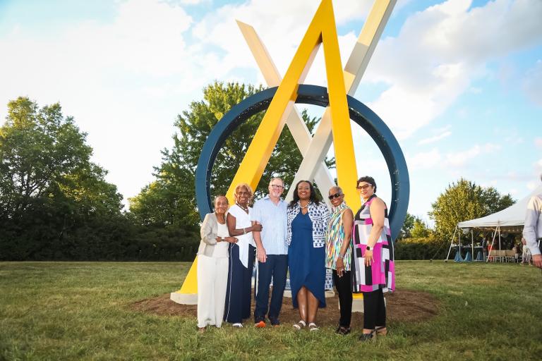 A group of people standing and smiling before a large yellow and blue geometric sculpture outdoors.