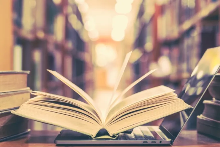 Open book on table with other books and laptop in front of a book case