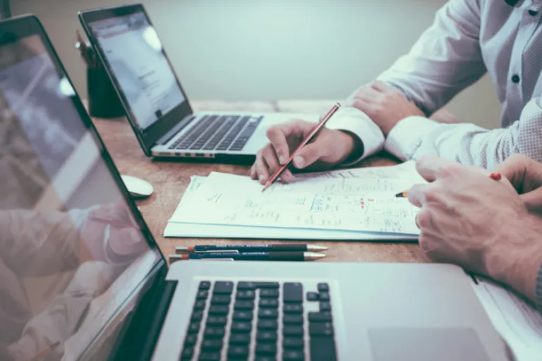 Two laptops and two people looking with both hands on stack of paper