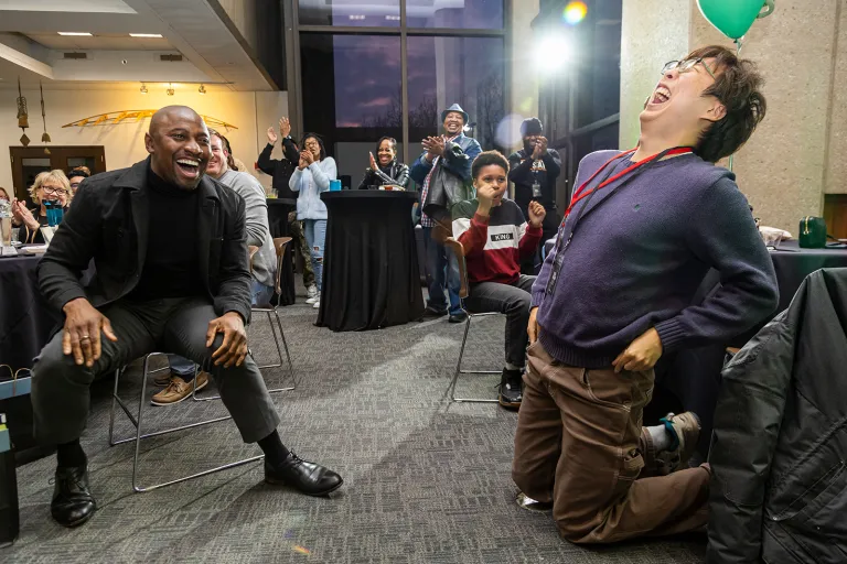 Two people laugh enthusiastically, one kneeling on the floor, surrounded by a cheering group of people at an indoor event.