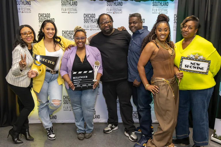 Seven people stand together smiling in front of a Chicago Southland Film Festival backdrop, holding film-themed props such as a director’s clapperboard and a “Now Showing” sign.