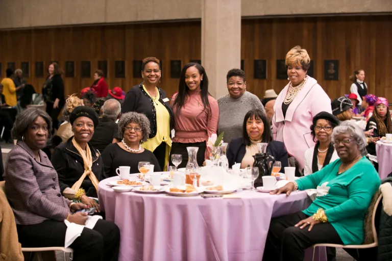 Group of people seated and standing around a table covered with a lavender tablecloth at a formal event with food, drinks, and table settings