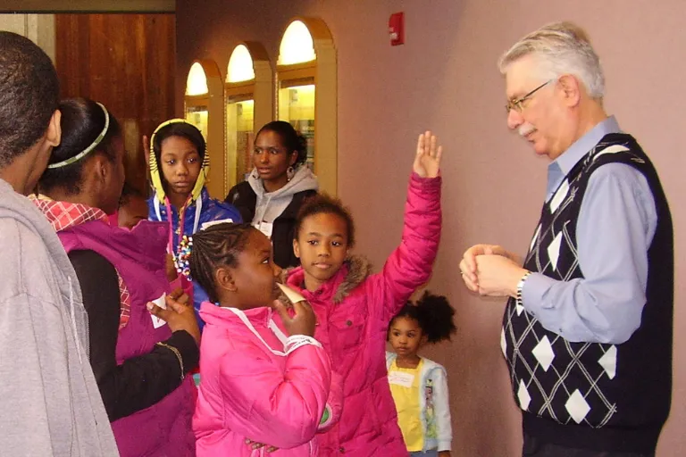 A group of children in winter coats gather around an adult in a hallway, with one child raising a hand as the adult speaks.