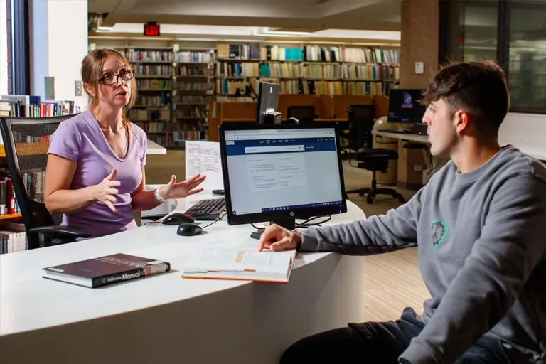Two people sitting at a table having a discussion, with one holding a tablet and the other working on a laptop in a library setting.
