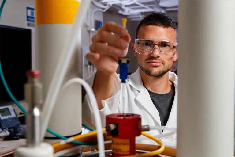 Scientist in lab coat and glasses examining a red liquid sample in a laboratory beaker.