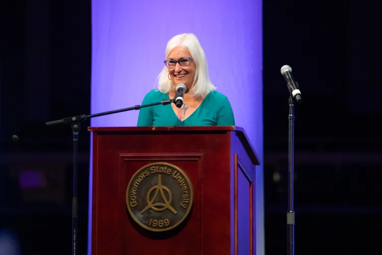 Smiling woman with white hair and glasses speaks at a podium with microphones.