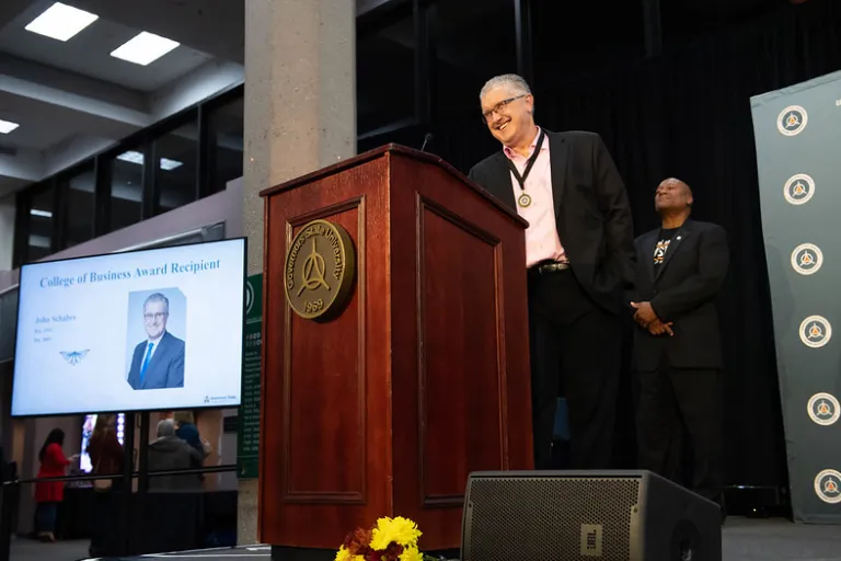 Smiling man in a suit speaks at a podium; screen behind him shows his award portrait.