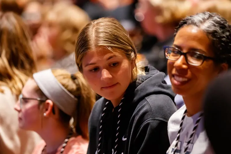 Attentive young woman in black hoodie next to a smiling woman with glasses among a crowd.