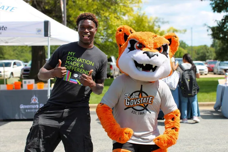 Student in t-shirt giving thumbs up next to a smiling tiger mascot outdoors.
