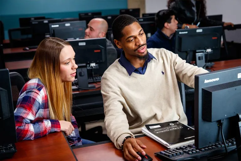 Man points at computer screen, assisting woman in a busy computer lab.