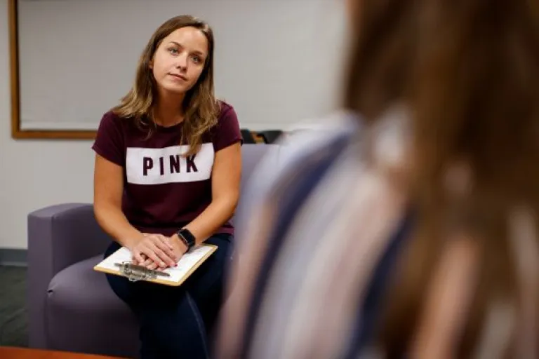 Woman with long brown hair, in a t-shirt, holding a clipboard, listening to someone out of frame.