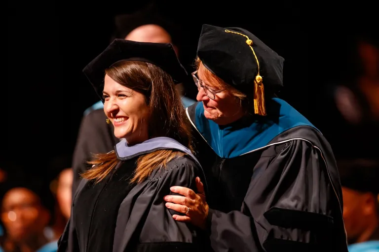 Doctoral graduate smiles as a faculty member places their hand on their shoulder during the Doctoral Hooding Ceremony.