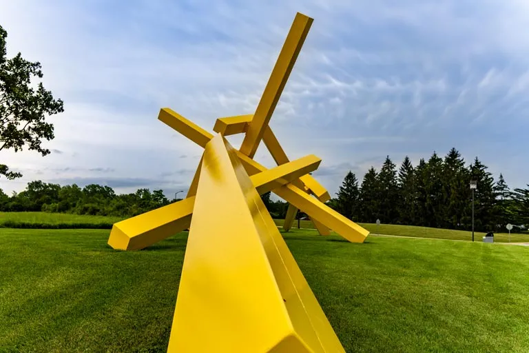 Abstract yellow beam sculpture in a park setting.