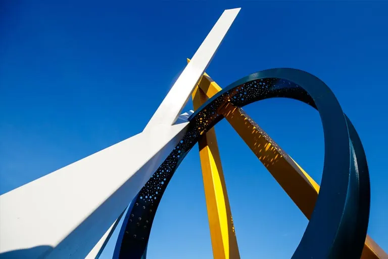 Abstract sculpture with angular white, curved yellow, and perforated blue elements against a clear sky.