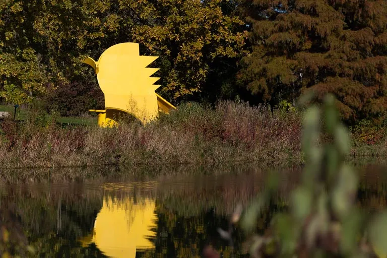 Large yellow bird sculpture by a pond, surrounded by fall trees.