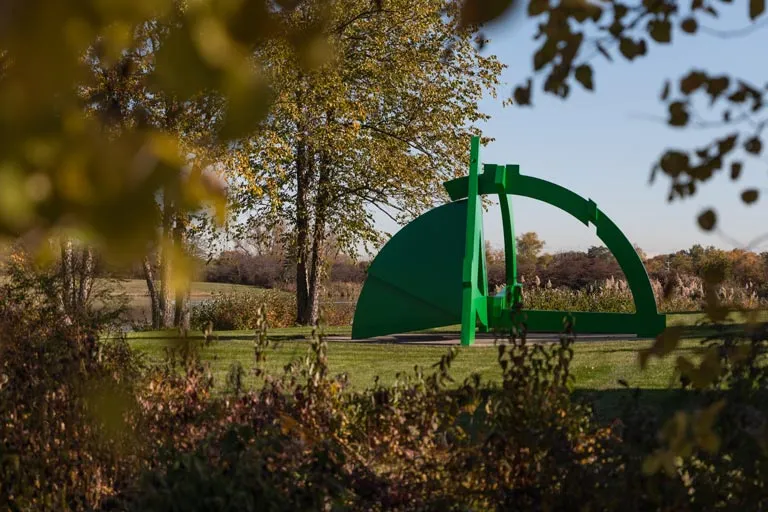 Bright green abstract sculpture on a grassy lawn, framed by autumn leaves.
