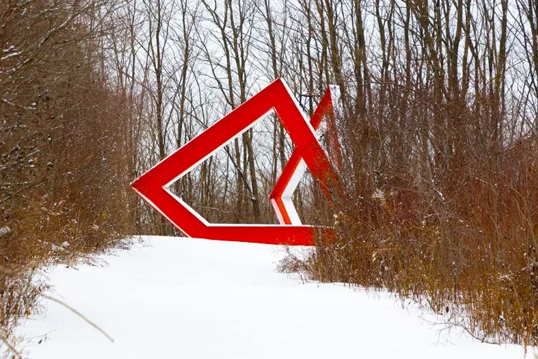 Abstract red sculpture on a snow-covered path, framed by bare trees.