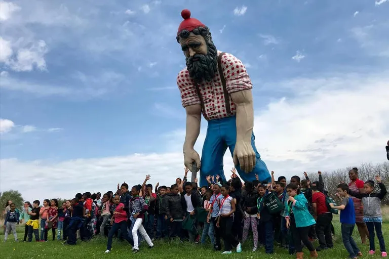 Giant lumberjack statue with a group of diverse children raising their hands in a grassy field.