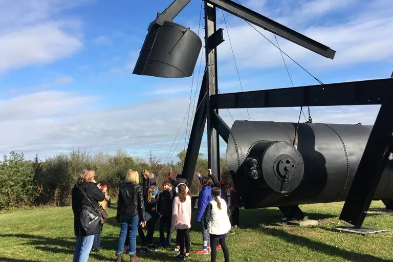 Group of children and adults viewing a large black industrial sculpture outdoors.