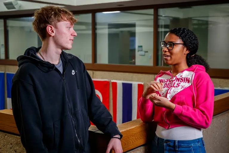 Young man in black hoodie and woman in pink hoodie talking in a bright hallway.
