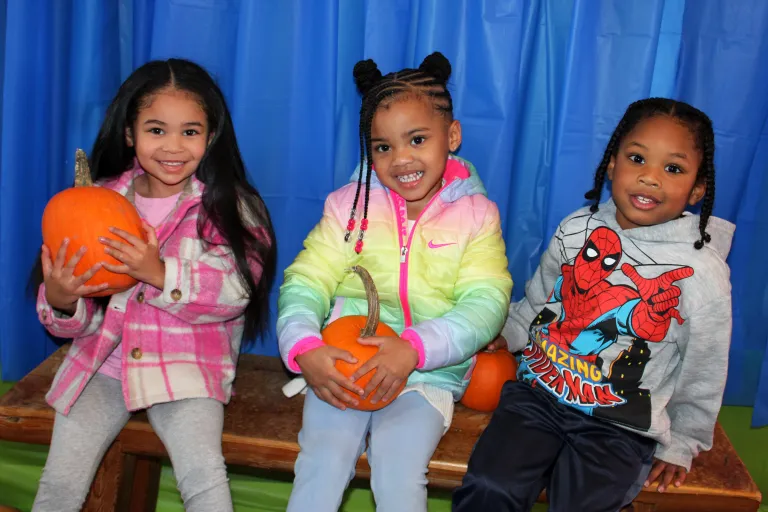 Three young children, sitting on a bench, holding pumpkins