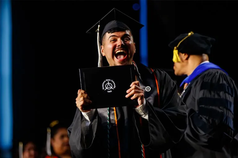 Excited male graduate in cap and gown holds diploma, mouth open in celebration.