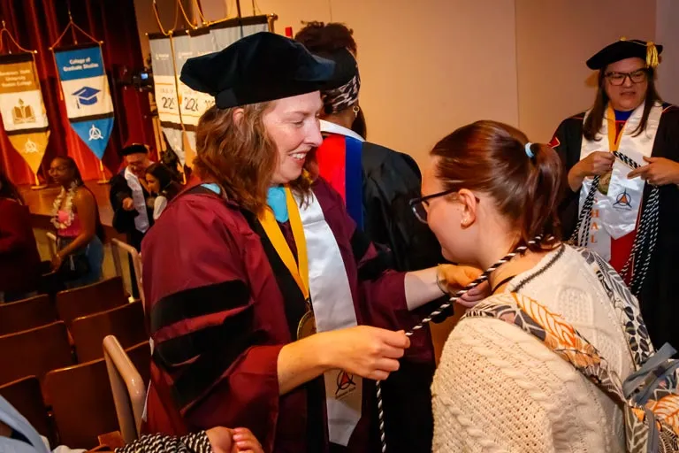 Smiling graduate in cap and gown places an honor cord on a student.
