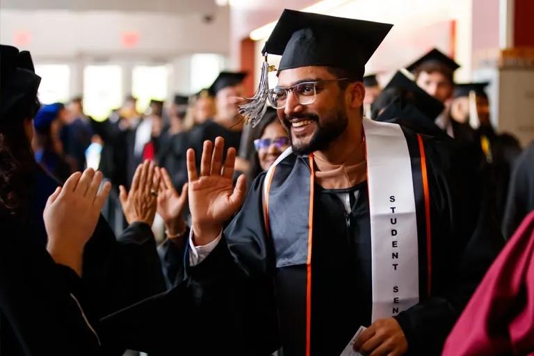 Smiling male graduate in cap and gown with "STUDENT SPEAKER" stole, waving.