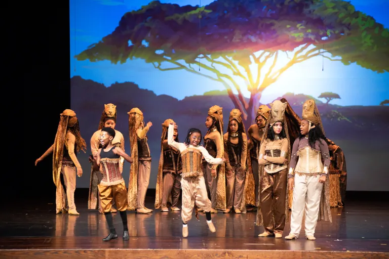 Theatrical performance with actors in animal costumes on stage, savanna backdrop.