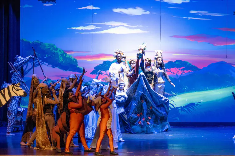 Vibrant stage show with actors in animal costumes and a savanna backdrop.