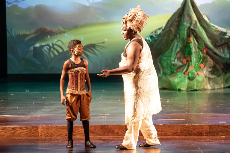 Teen performer in ornate costume gestures to child in earthy attire and mask on stage.