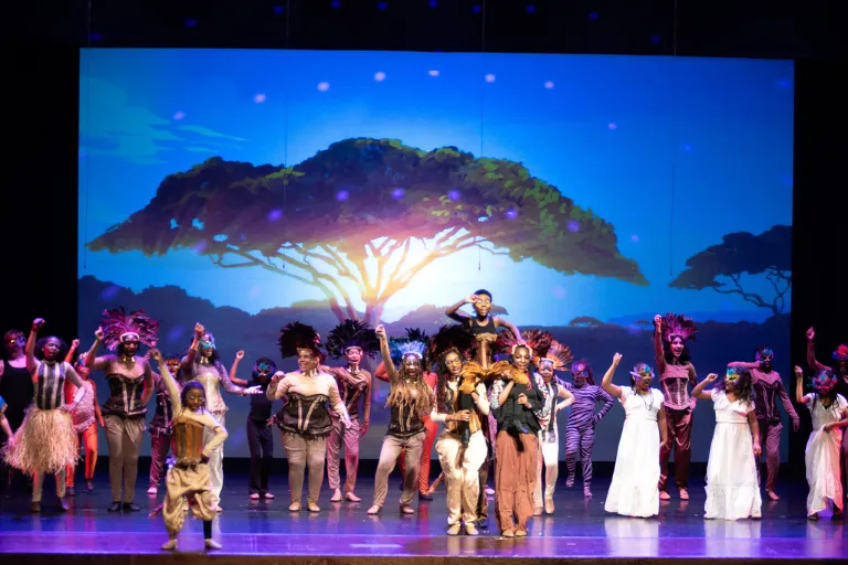 Stage performance with many actors in elaborate costumes against a tree silhouette backdrop.