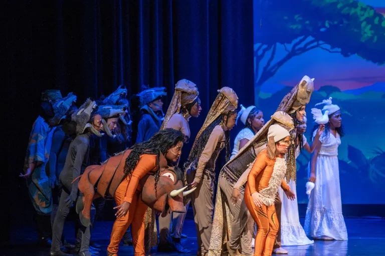 Children in various animal costumes walk left across a stage with a savanna backdrop.