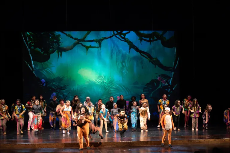 Group of performers in bright costumes on stage, glowing jungle scene projected behind them.
