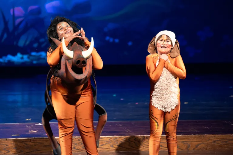 Two actors in Pumbaa and Timon costumes smiling on a dark stage.
