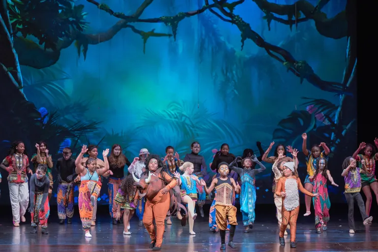 Kids in vibrant jungle-themed costumes dance on stage with a blue-green forest backdrop.