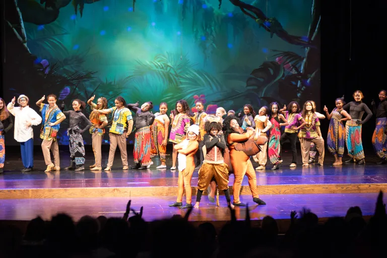 Performers in colorful costumes on a stage with a jungle backdrop, audience in foreground.