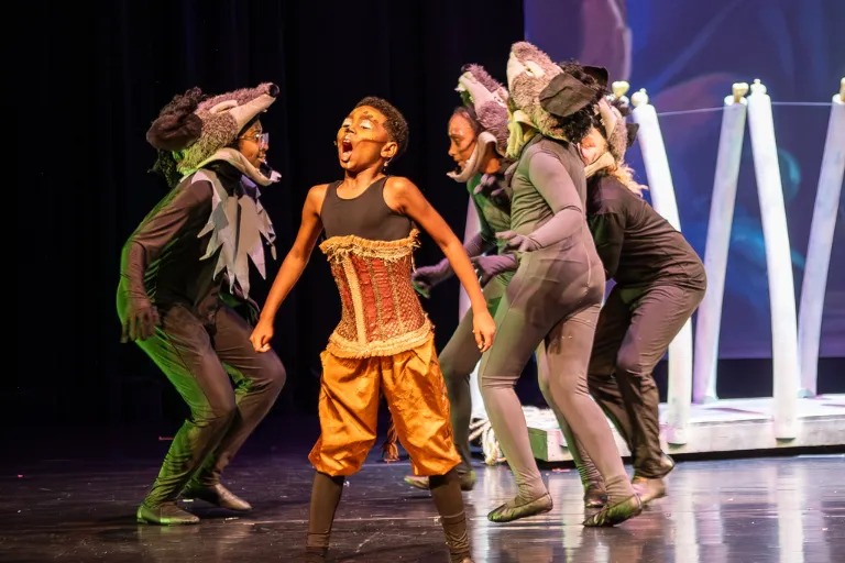 Child in orange and corset singing dramatically on stage, surrounded by performers in hyena costumes.