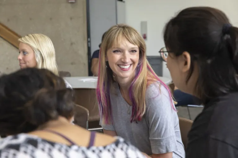 Smiling woman with long blonde and pink hair, sitting at table, talking with others
