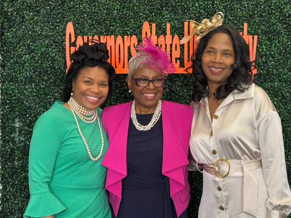 Three smiling women in formal attire and hats, standing before a green "Governors State University" wall.