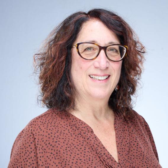 Smiling woman with dark curly hair and tortoise-shell glasses, wearing a brown top.