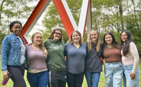 Group of seven female students, standing in front of large outdoor sculpture. 