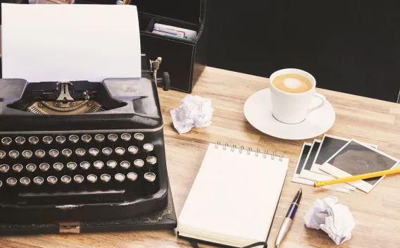 Typewriter next to messy desk of notepad, coffee, and crumpled papers