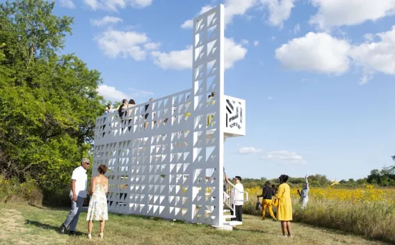 White geometric art installation with people in a sunny, flowery field.