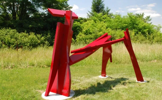 Abstract, bright red metal sculpture on a sunny green lawn with foliage.