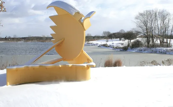 Golden abstract sculpture in snow next to a partially frozen lake under a cloudy sky.